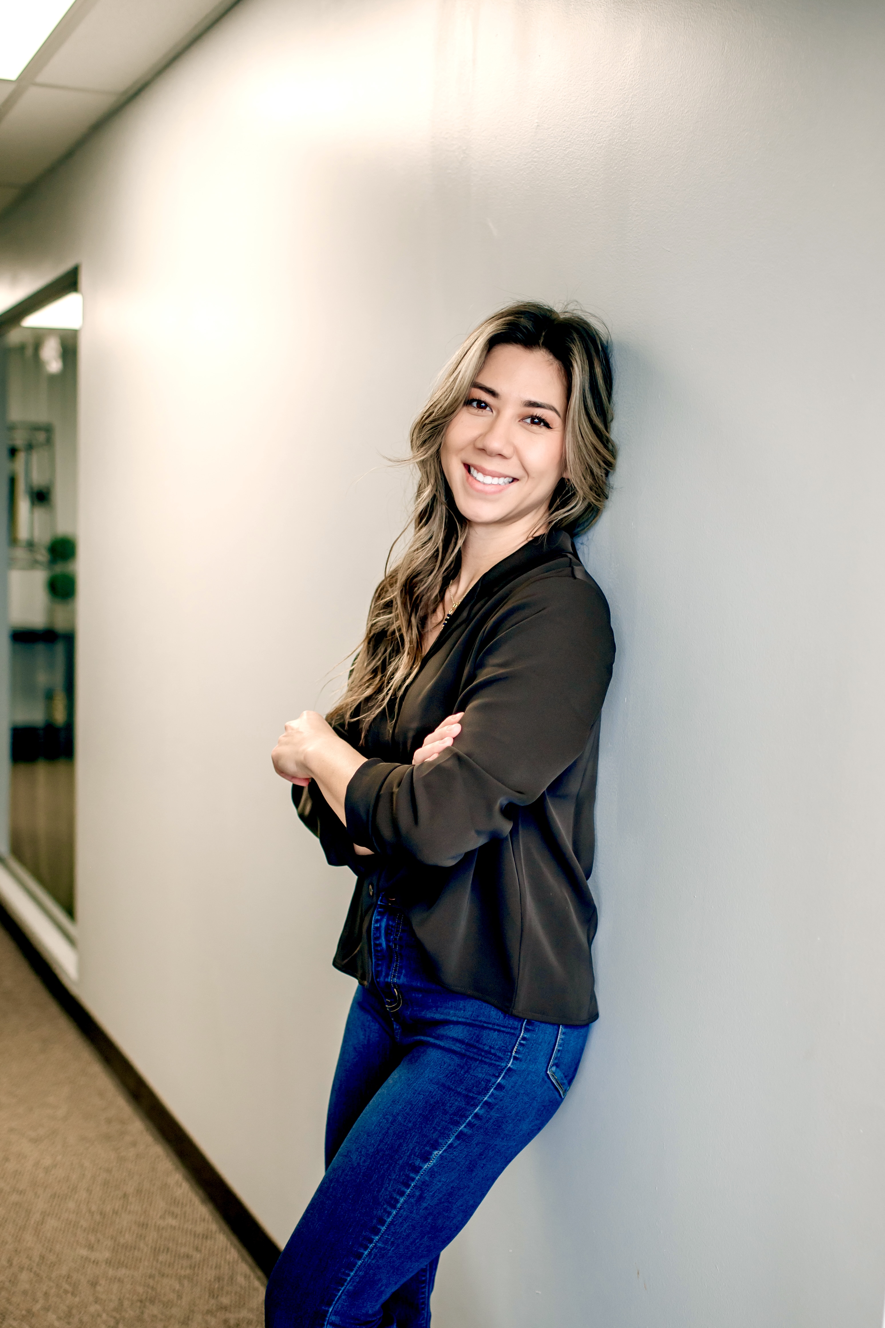 Smiling woman with long wavy hair wearing a black blouse and blue jeans leaning against a light gray wall in a hallway.