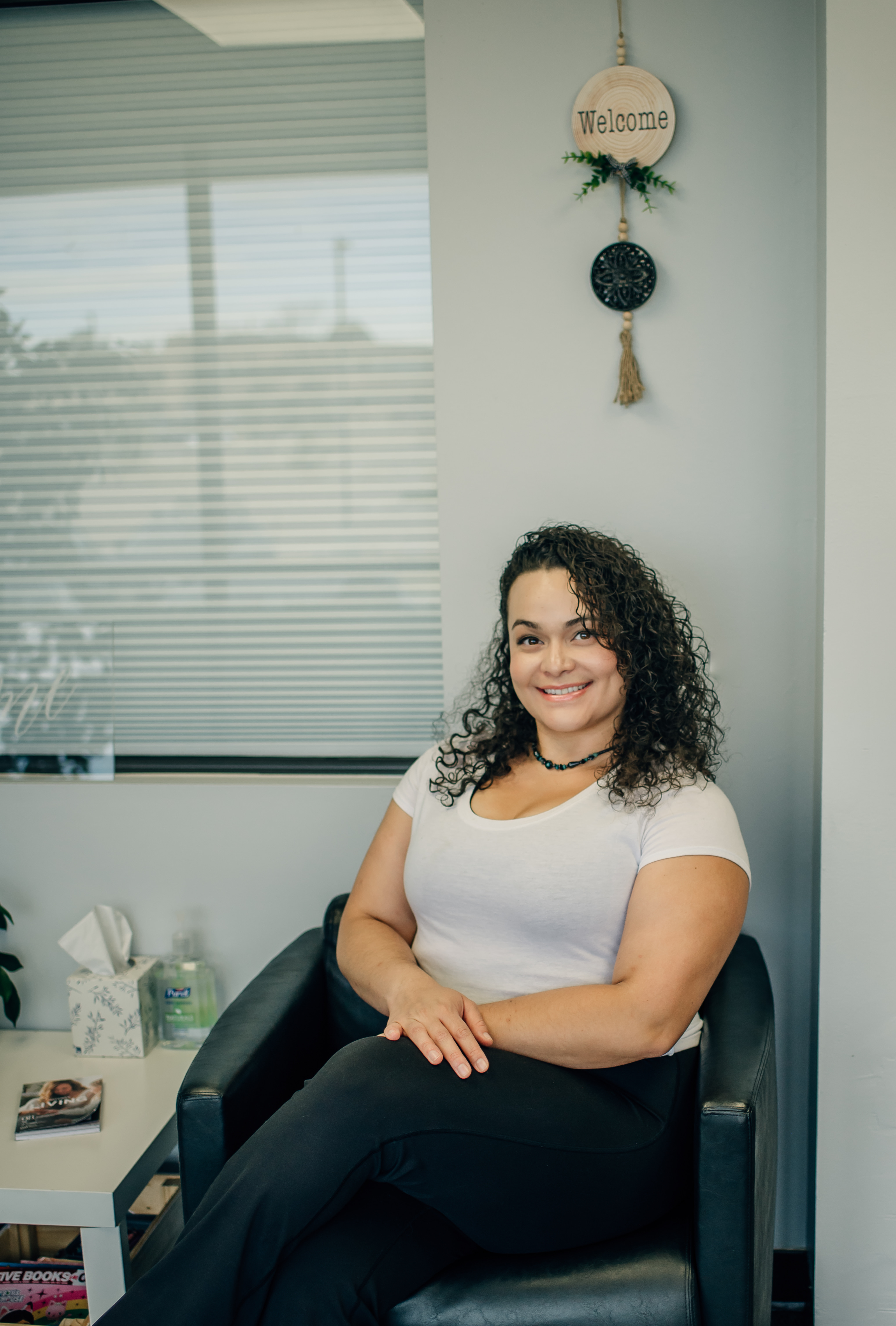 Smiling woman with curly hair wearing a white shirt and black pants sitting on a black chair in a modern room.