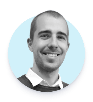 Retrato de un hombre sonriente con camisa blanca y suéter oscuro, fondo azul claro.