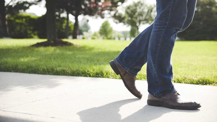 Person wearing brown shoes and blue jeans walking on a sunny sidewalk next to green grass.