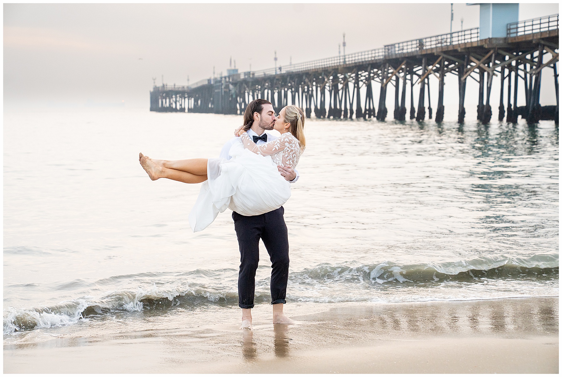 Couple running along Seal Beach during a December engagement session. Bride-to-be wearing a flowing white dress and groom-to-be dressed in a black suit with bow tie. Wind catching the dress as they splash through the shoreline