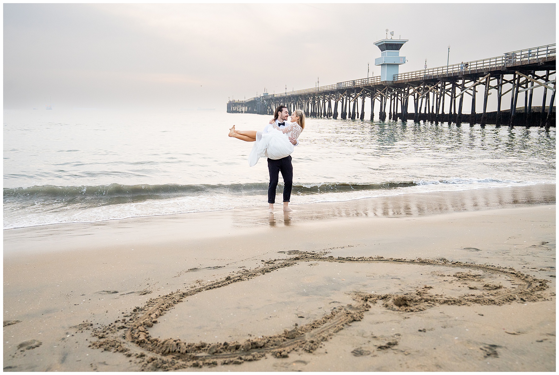 Couple running along Seal Beach during a December engagement session. Bride-to-be wearing a flowing white dress and groom-to-be dressed in a black suit with bow tie. Wind catching the dress as they splash through the shoreline