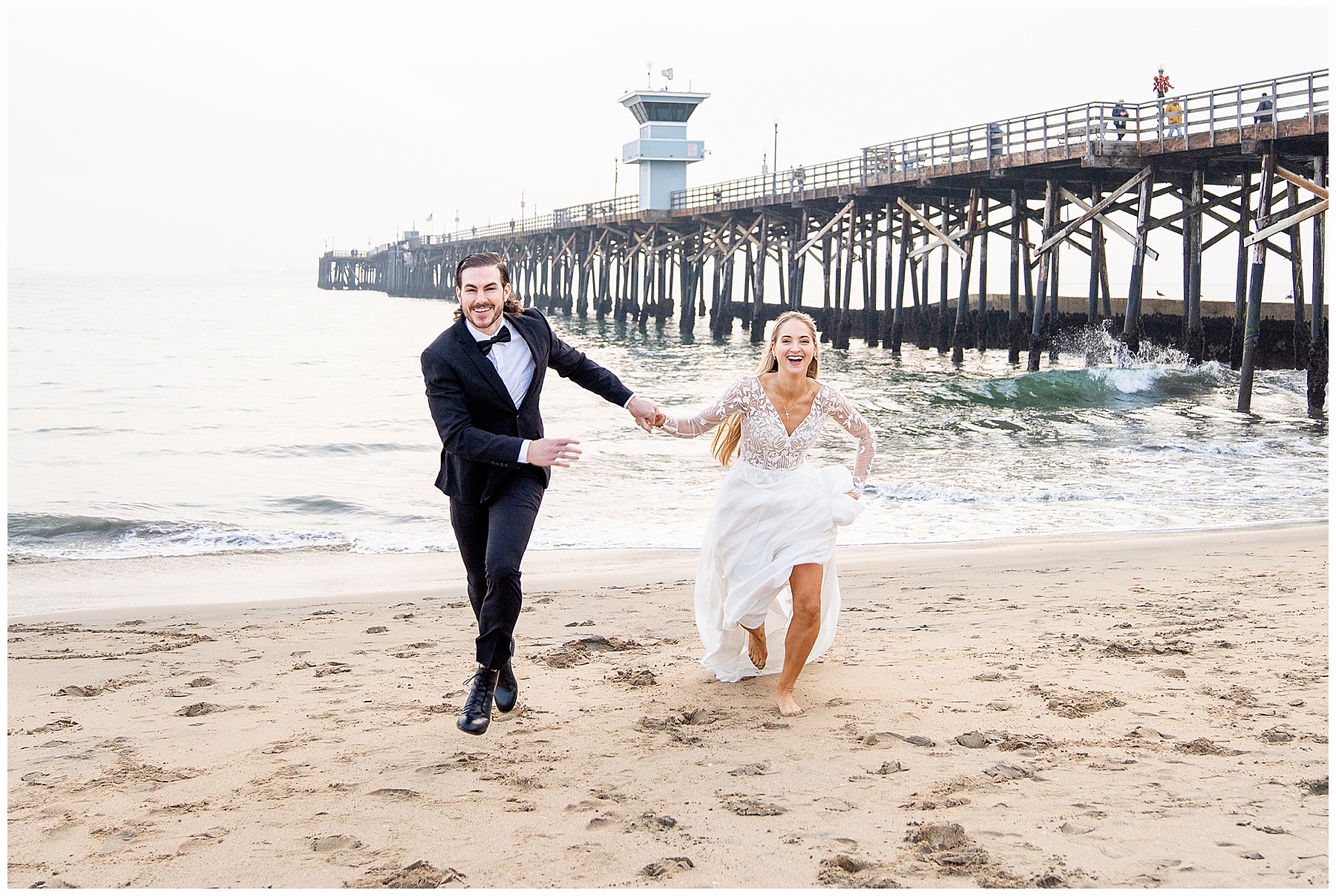 Couple running along Seal Beach during a December engagement session. Bride-to-be wearing a flowing white dress and groom-to-be dressed in a black suit with bow tie. Wind catching the dress as they splash through the shoreline