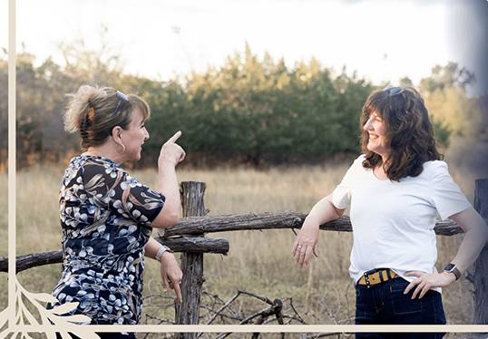 Two women smiling and talking while leaning on a rustic wooden fence in a natural outdoor setting.