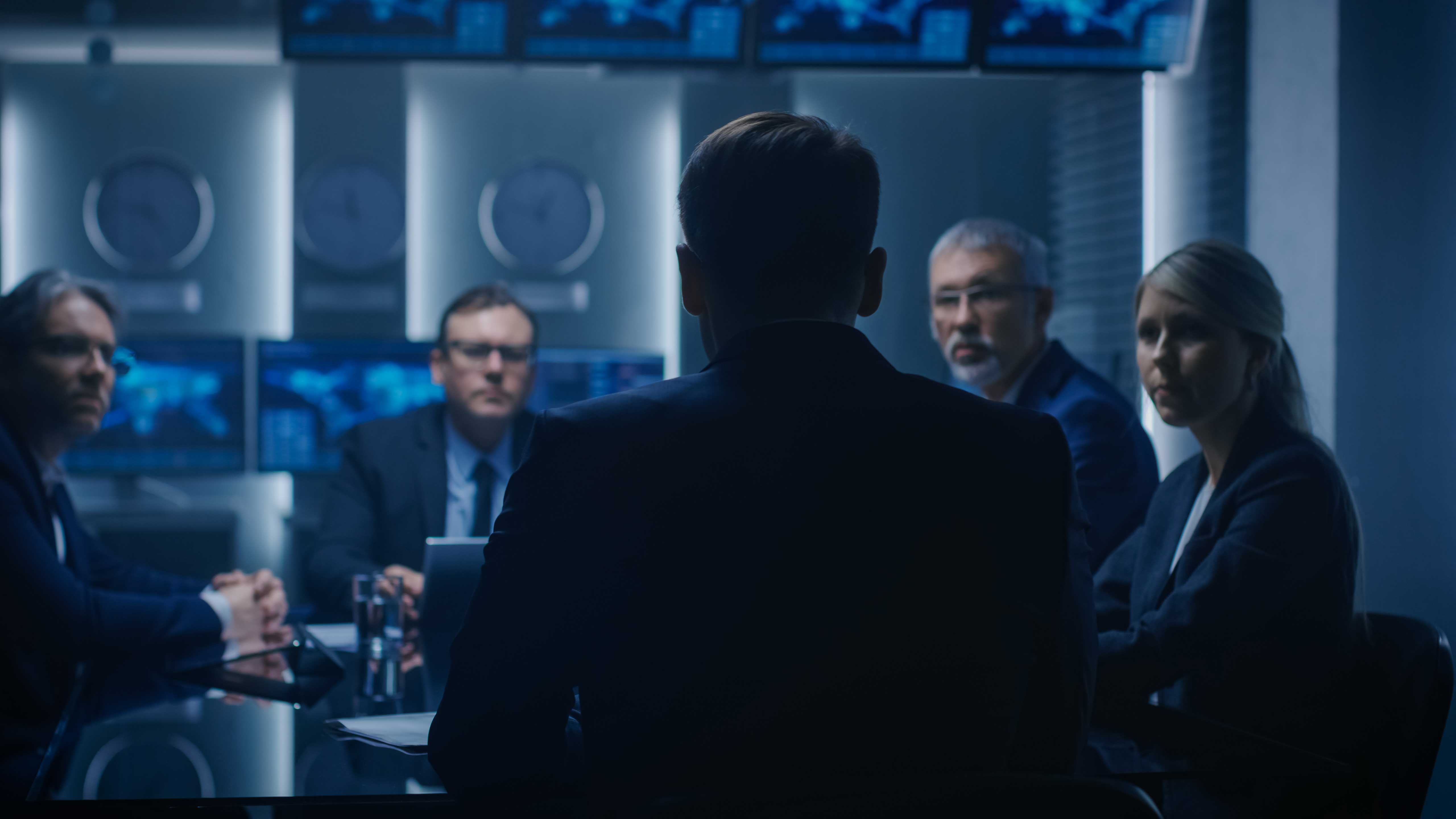 Secret meeting in a high-tech conference room with a man seen from behind and four colleagues facing him.