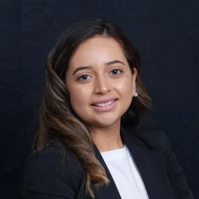 Smiling woman with long, wavy brown hair wearing a black blazer and white top against a dark background.