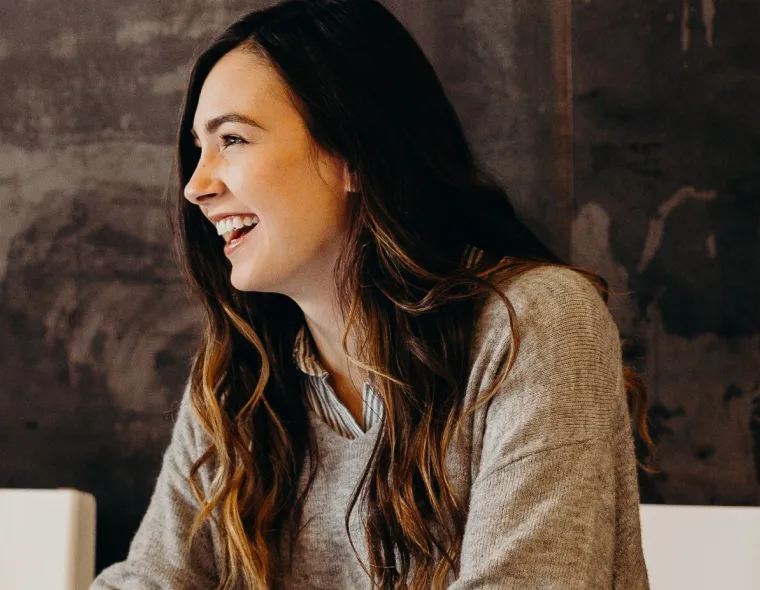 Young woman with long brown hair laughing and looking to the left against a dark textured wall.