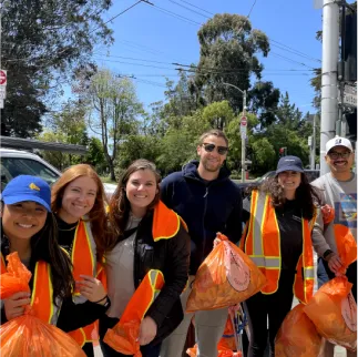 Group of six people outdoors wearing safety vests and holding orange bags, smiling at the camera.