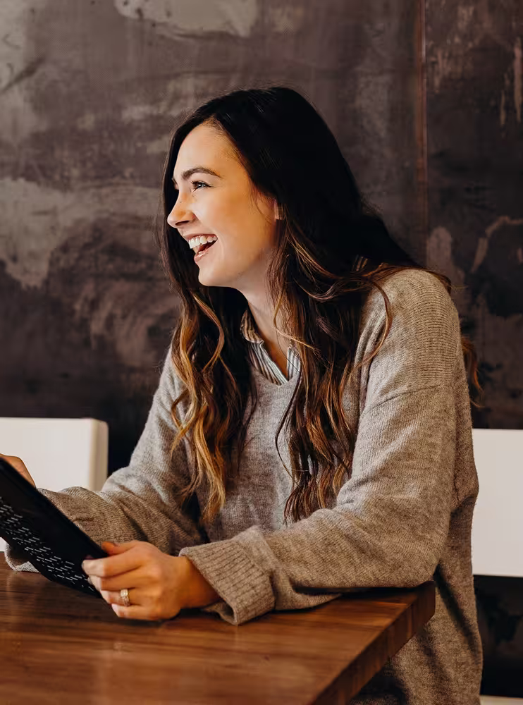 Smiling woman with long brown hair wearing a gray sweater, sitting at a wooden table holding a tablet.