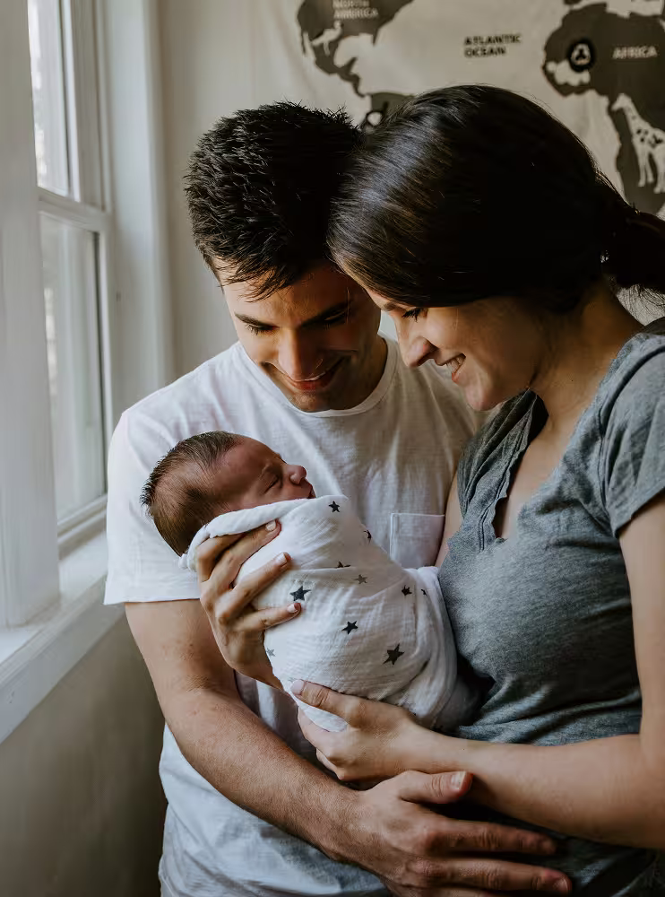 Smiling parents holding their newborn baby wrapped in a white blanket with stars near a window.