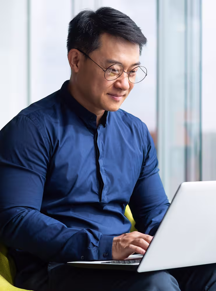 Man wearing glasses and a blue shirt typing on a laptop while seated near a window.