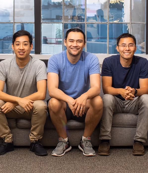 Three men sitting on a gray couch in front of window panes, smiling and facing the camera.