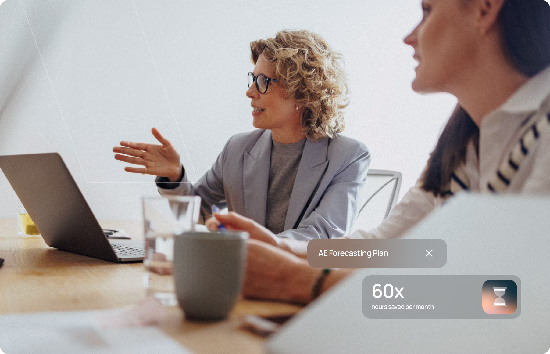 Two women in a meeting, one with curly blonde hair speaking and gesturing towards a laptop on the table.
