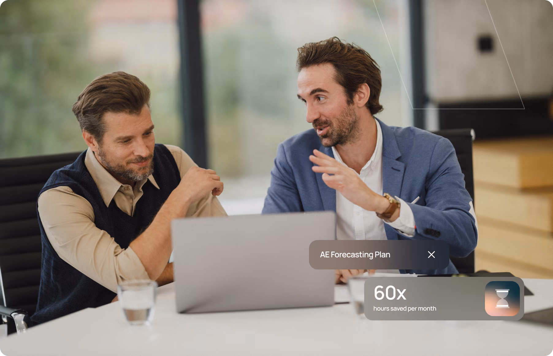 Two men discussing at a table with a laptop and a glass of water in a modern office setting.