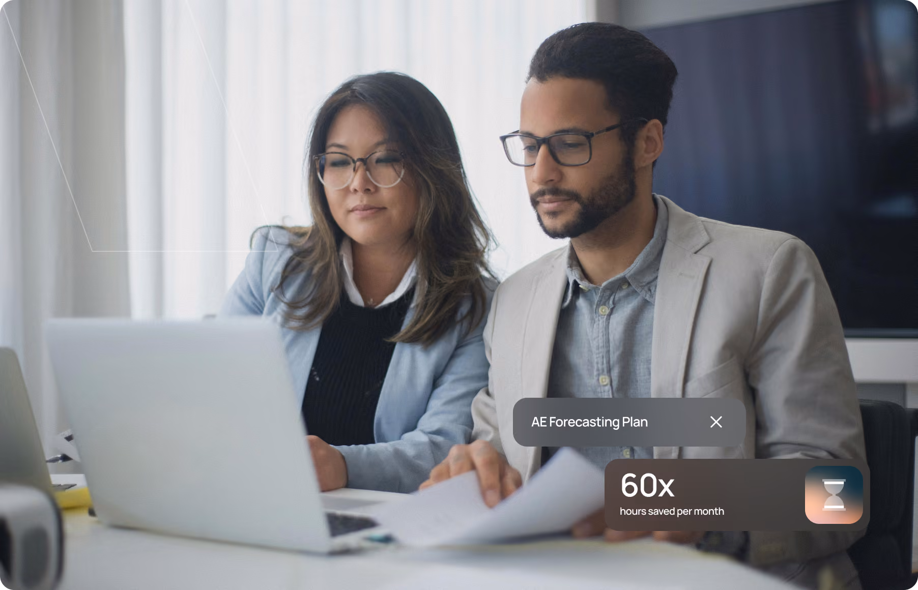 Two professionals wearing glasses review documents together at a laptop in a bright office.