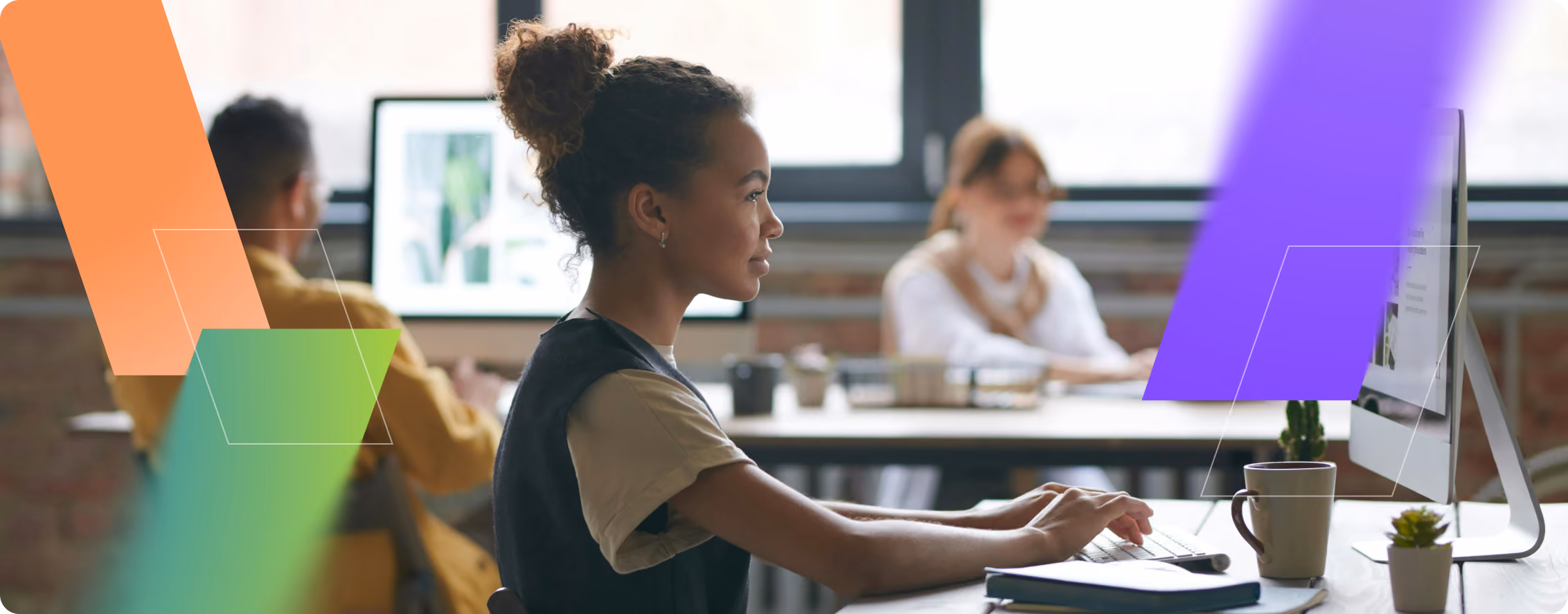 Young woman working at a desktop computer in a modern office with colleagues in the background.