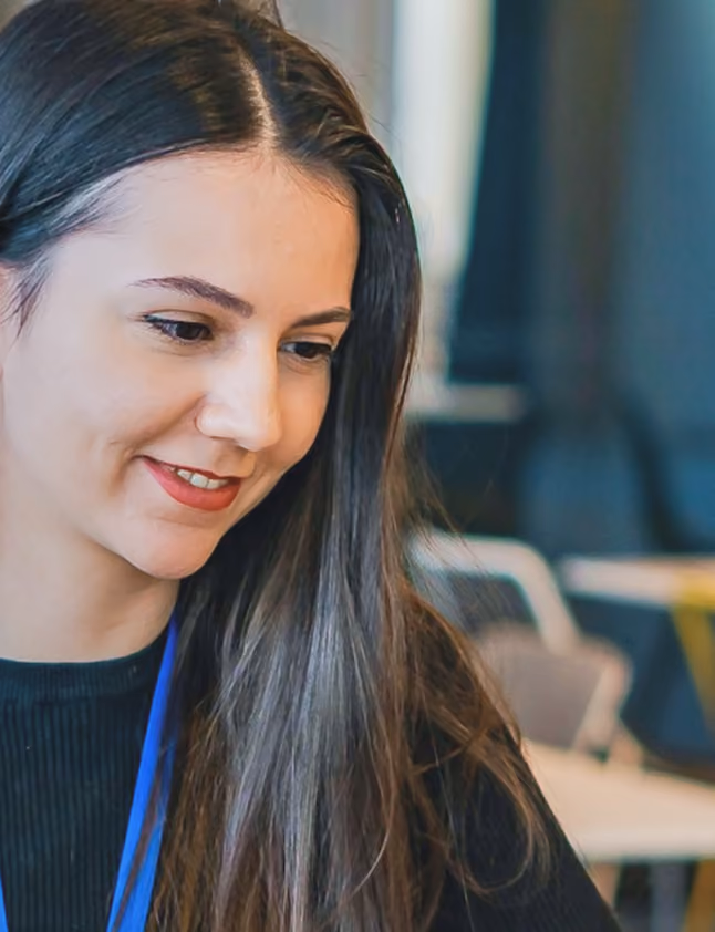 Smiling young woman with long dark hair wearing a black top and blue lanyard, seated indoors.