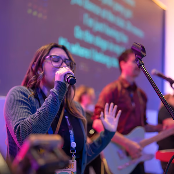 Woman singing into a microphone with musicians playing guitar in the background on stage.