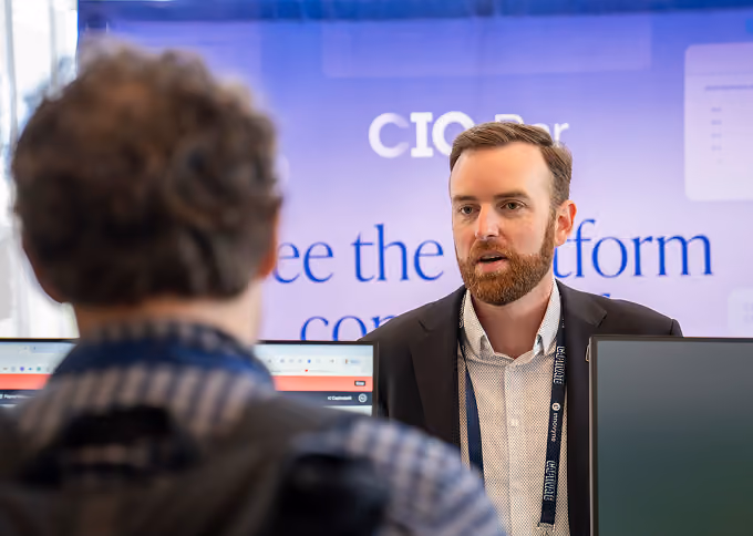 Man with beard and lanyard talking to another man in front of a booth with a blurred background.