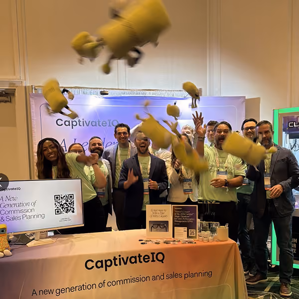 Group of people at CaptivateIQ booth joyfully tossing yellow plush toys in the air.