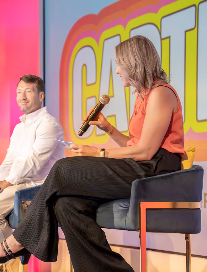 Woman in coral top holding a microphone seated next to a man in white on stage with colorful background.