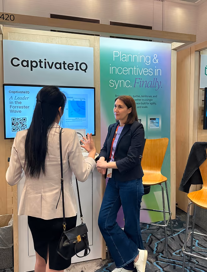 Two women talk at a CaptivateIQ booth with signs about planning and incentives synchronization.