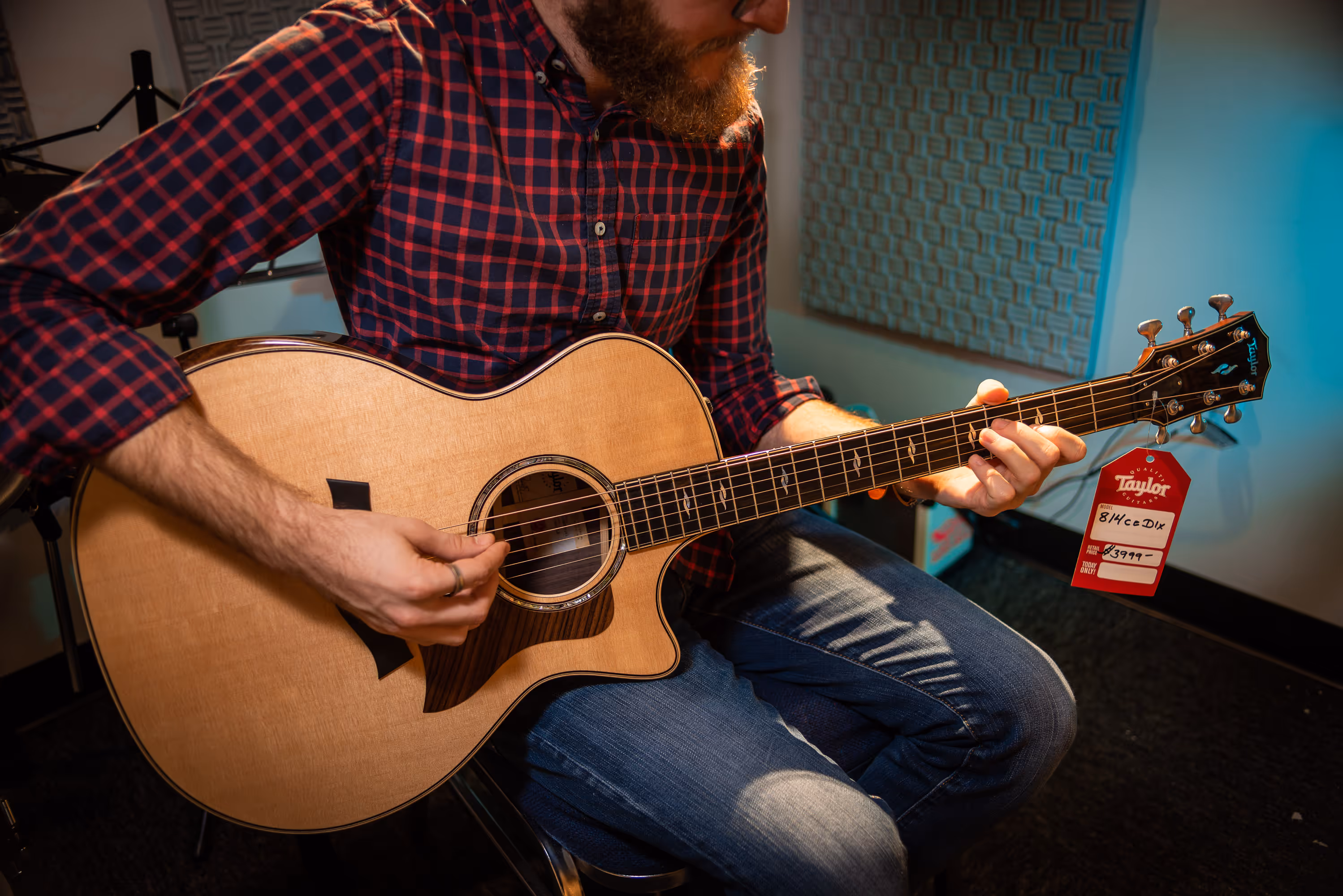 Man in red and black checkered shirt playing a Taylor acoustic guitar with price tag attached.