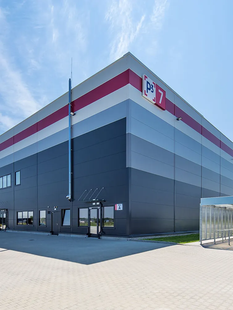 Modern warehouse building with horizontal stripes in red, light blue, and dark gray under a clear blue sky.