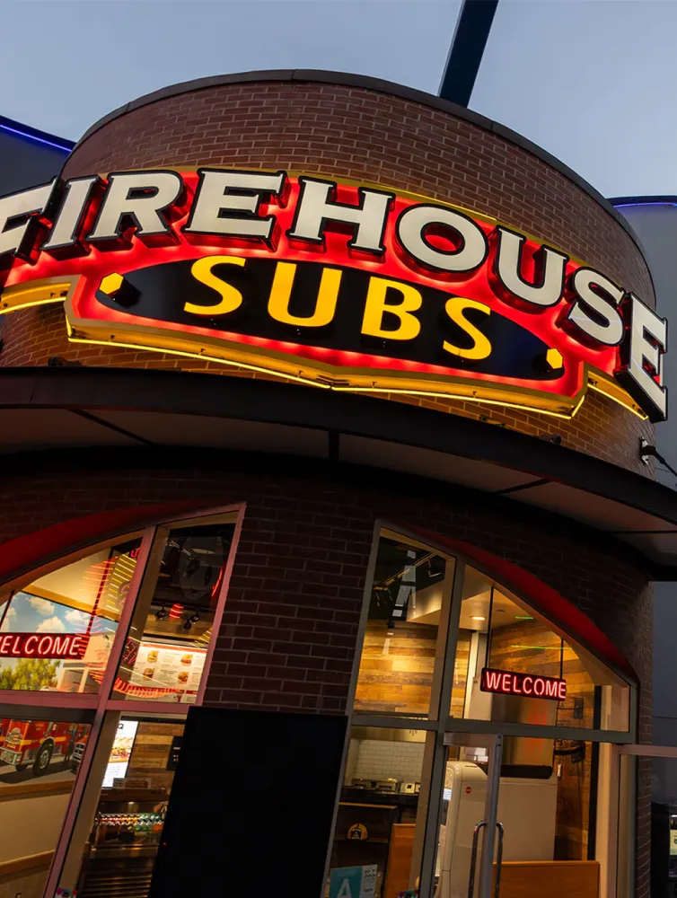 Exterior of Firehouse Subs restaurant with illuminated red and yellow sign and glass entrance displaying a welcome neon sign.
