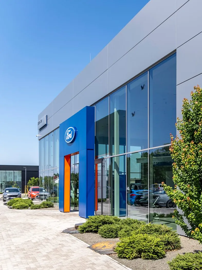 Modern Ford dealership building with large glass windows, blue and orange entrance, and cars parked outside on a sunny day.
