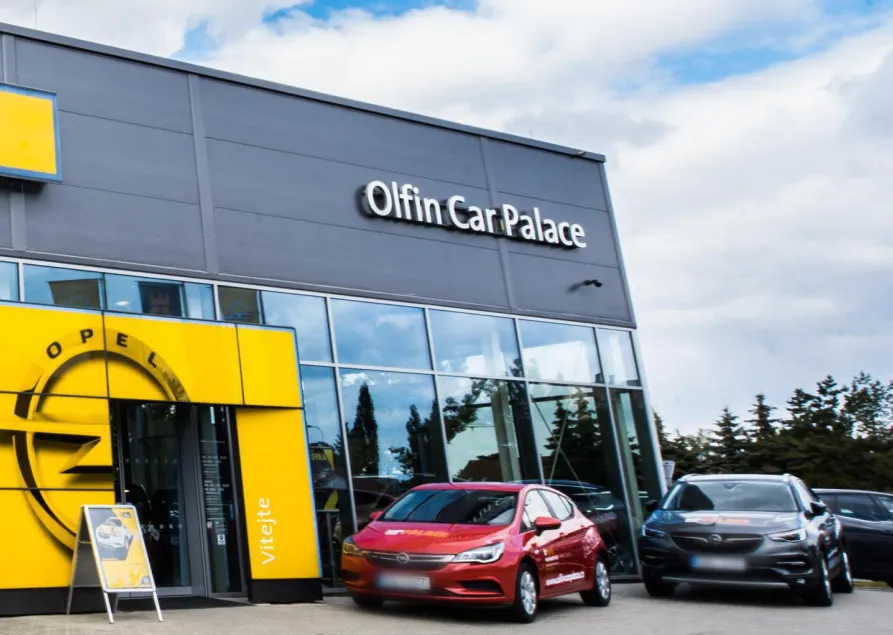 Exterior of Olfin Car Palace dealership with two cars parked in front, one red and one black, under a partly cloudy sky.