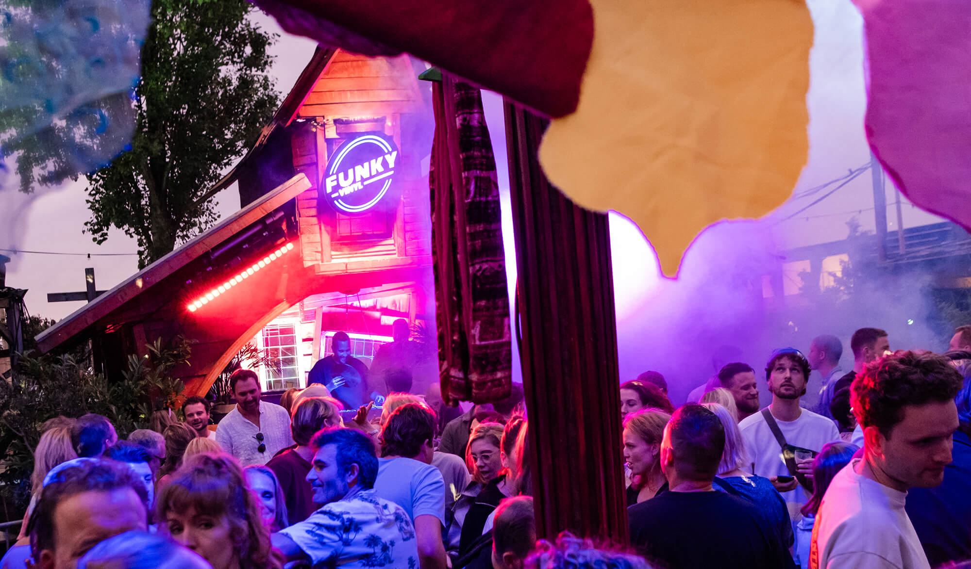 Crowd of people enjoying an outdoor party with colorful lights and a DJ booth under a sign reading 'FUNKY VINYL'.