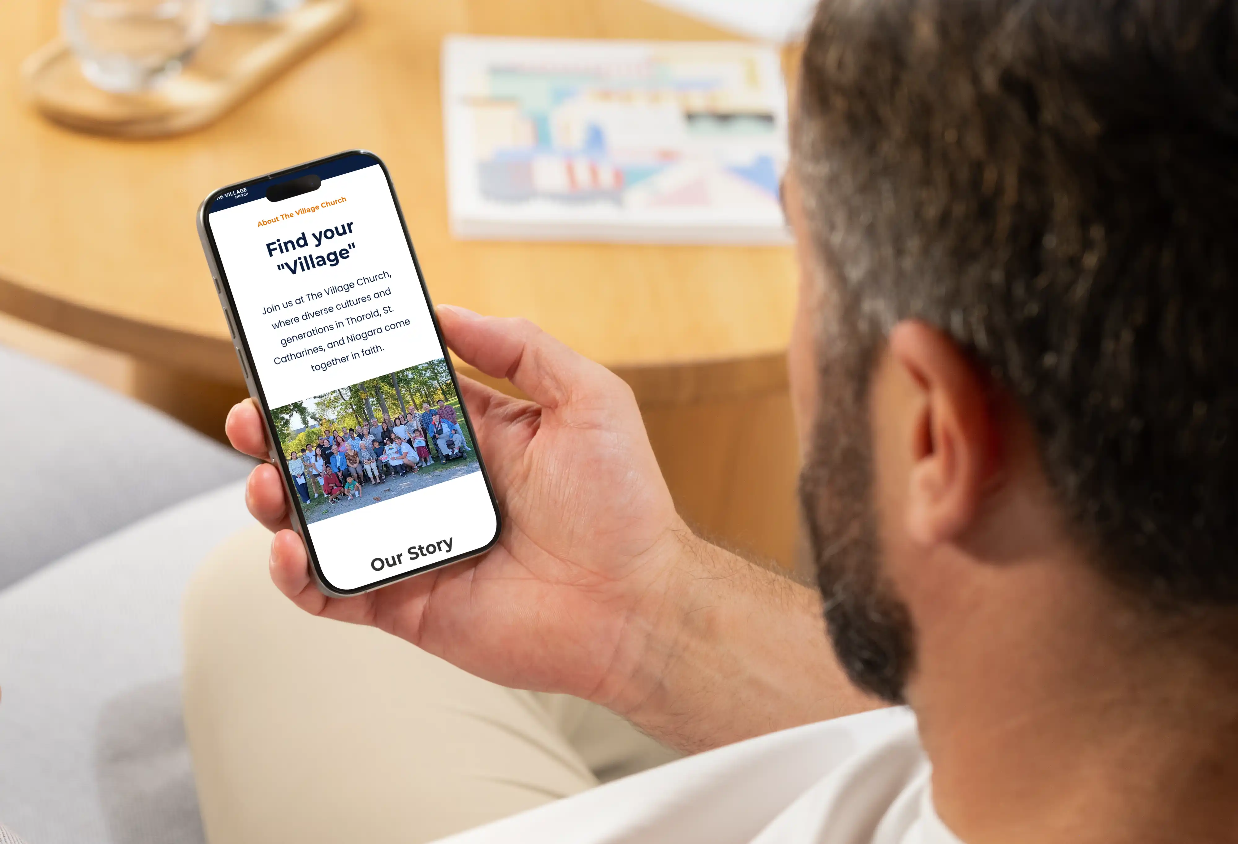 A man holds a smartphone displaying the "About Us" page of The Village Church website redesign.