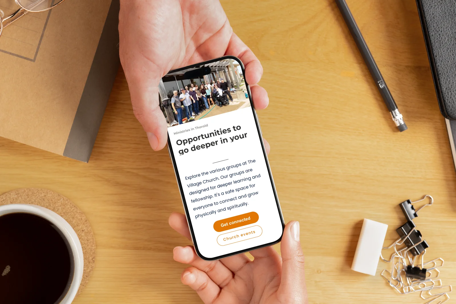 A person passes a smartphone displaying the "Ministries" CTA section of The Village Church website redesign to another person. On the wooden desk are glasses, a notebook, pencil, binder clips, and a cup of coffee.