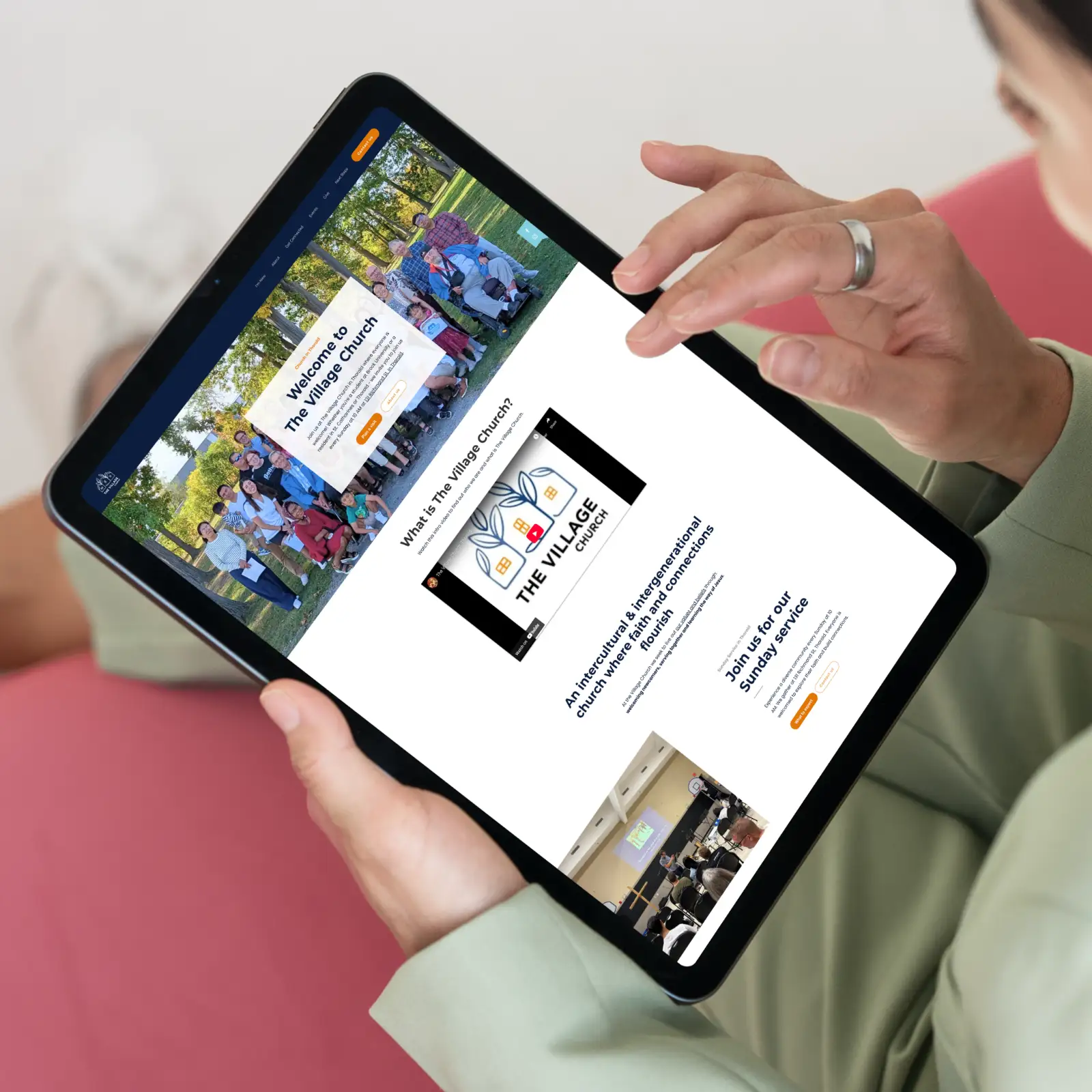 A person holds a tablet displaying The Village Church website redesign. The screen shows images of a diverse group and church information. The mood is welcoming and community-focused.