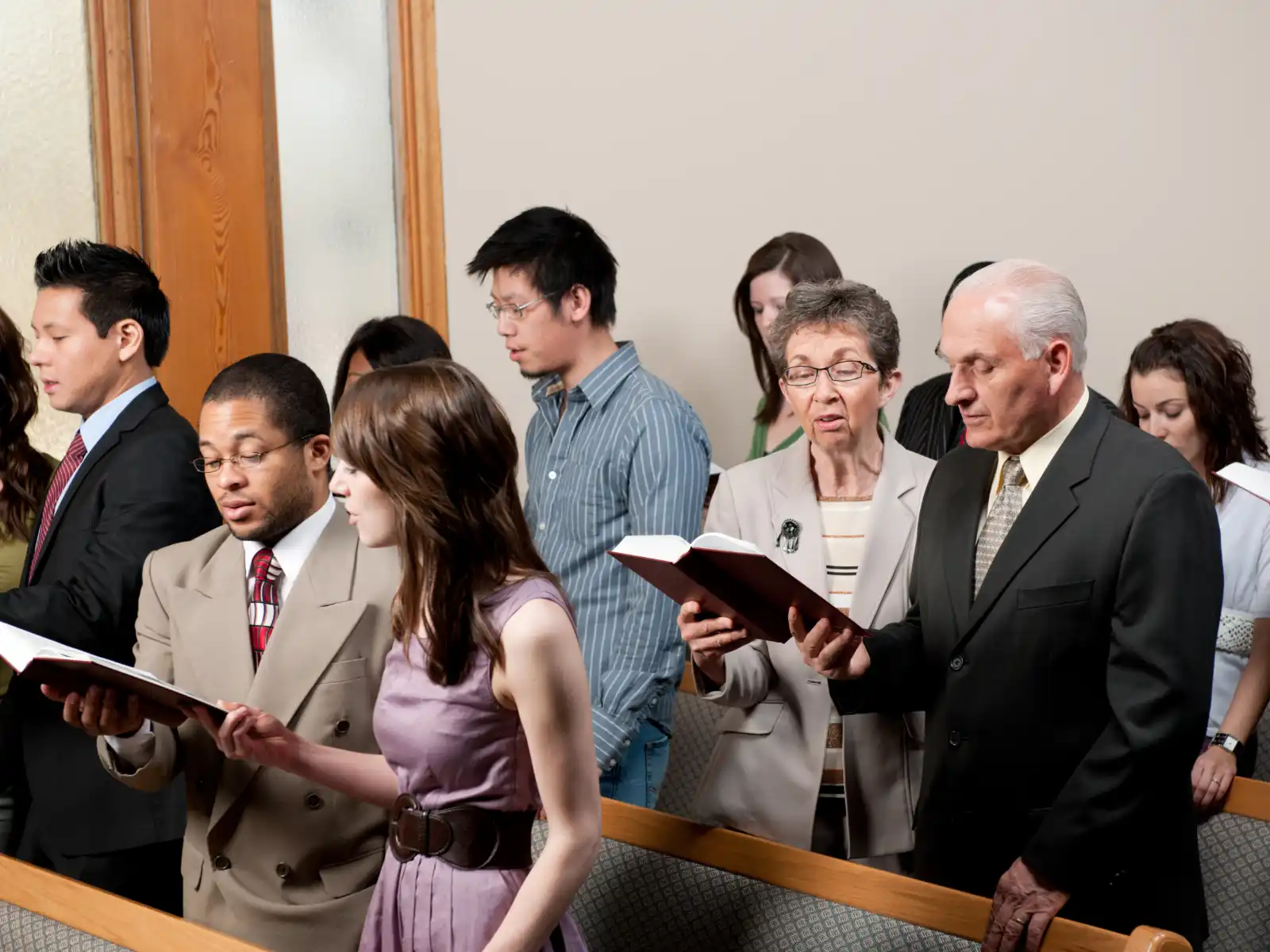A diverse group of people stand in a church, holding hymnals. They appear focused and engaged, suggesting reverence and unity during worship.