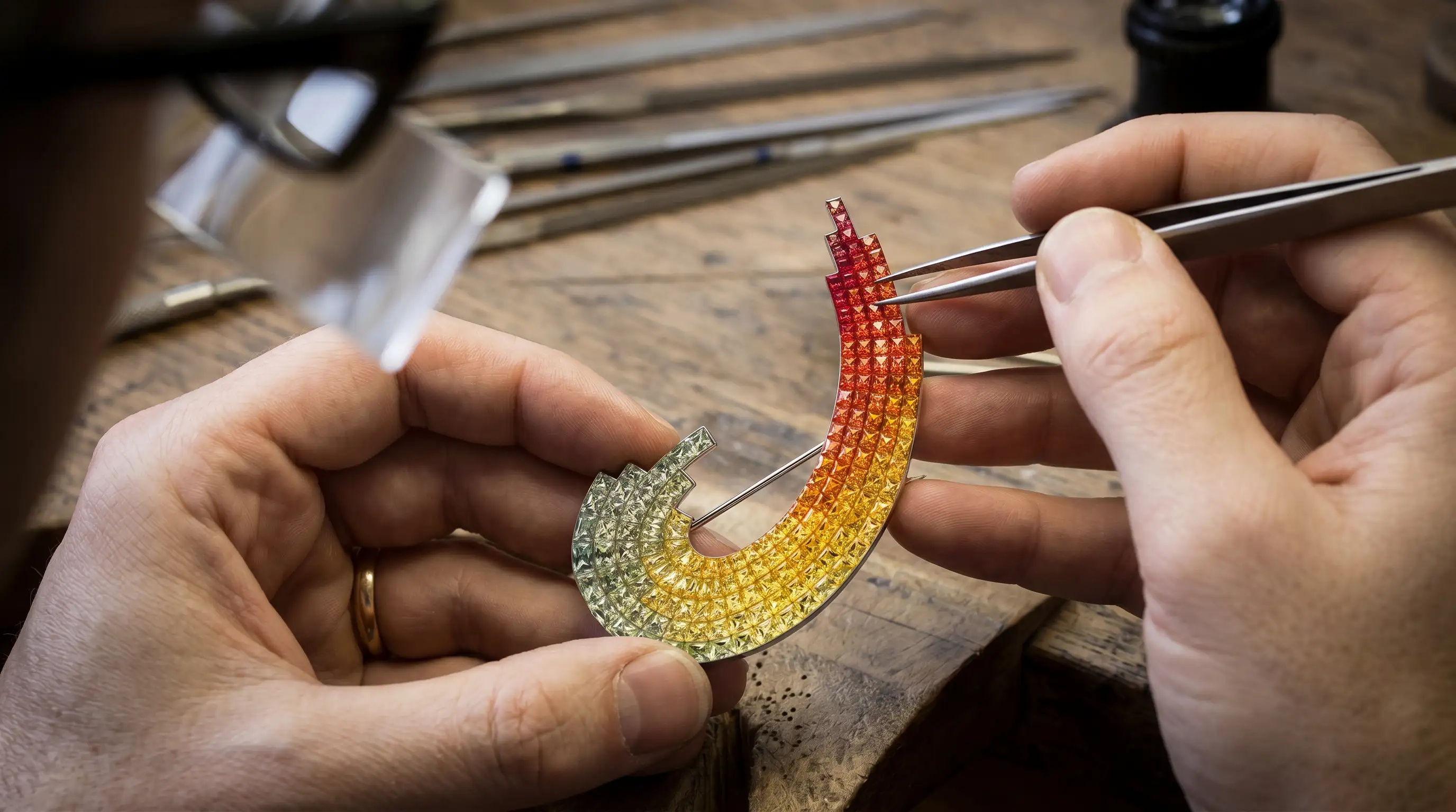 A professional jeweler using tweezers to assemble a custom gradient gemstone brooch featuring intricate pave-set red, orange, and yellow sapphires.