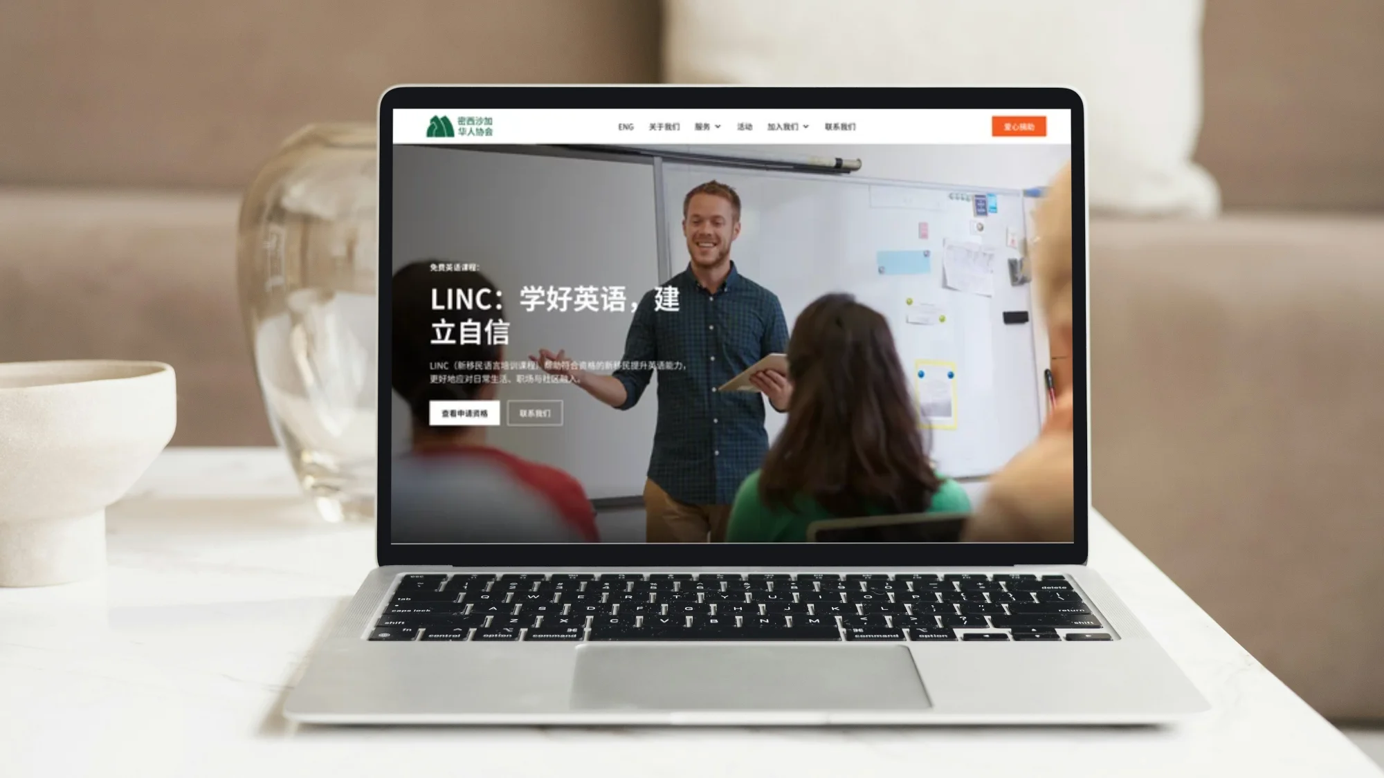 Open laptop on a table displaying the Chinese Association of Mississauga website with a man teaching in a classroom to students, alongside Chinese text promoting an English learning program called LINC.