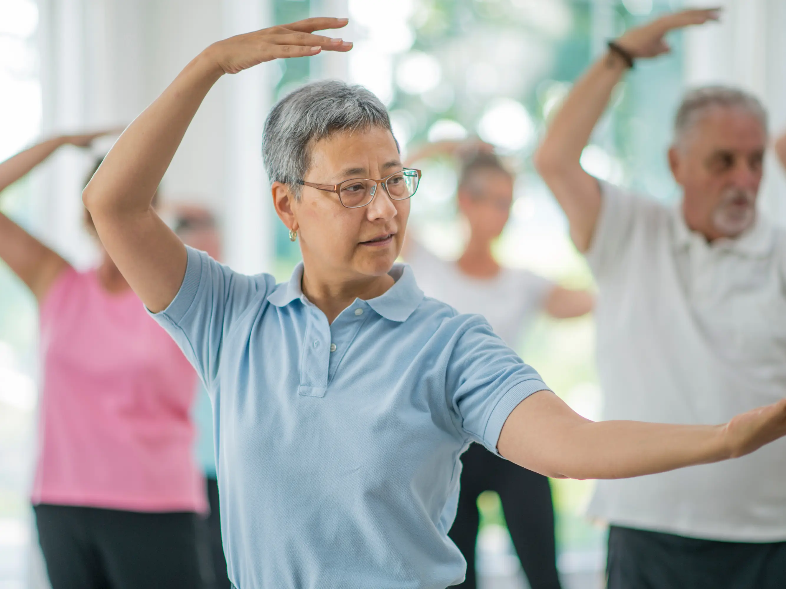 A group of older adults practicing tai chi indoors, with the woman in front wearing glasses and a light blue polo shirt raising her arms in a tai chi pose.