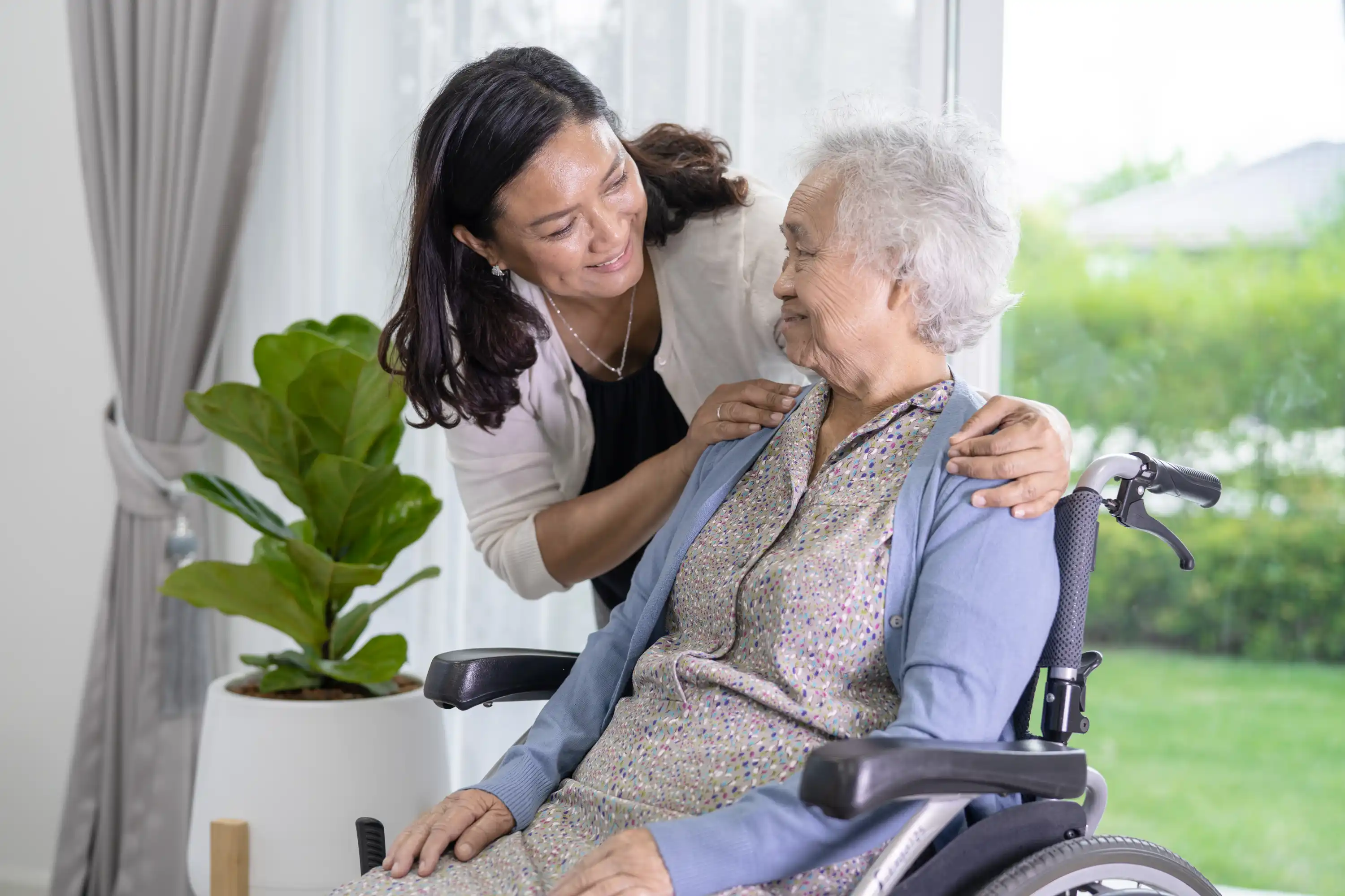 Smiling middle-aged woman gently embraces and talks to an elderly woman seated in a wheelchair inside a well-lit room with a large window and green plants outside.
