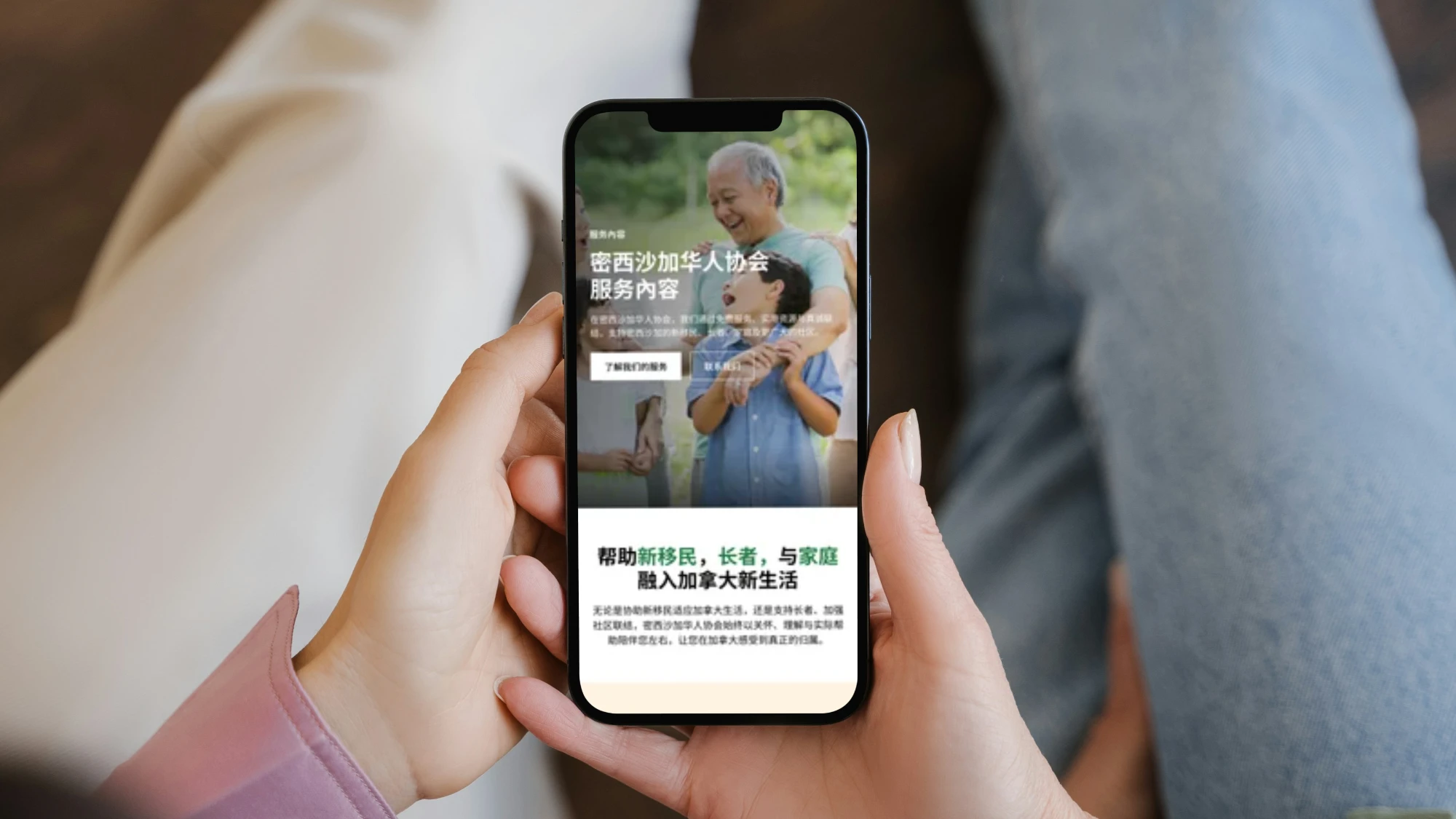 A person holding a smartphone displaying Chinese Association of Mississauga website in Chinese featuring an older man and a young boy smiling and hugging outdoors, with text about services for new immigrants, seniors, and families integrating into Canadian life.