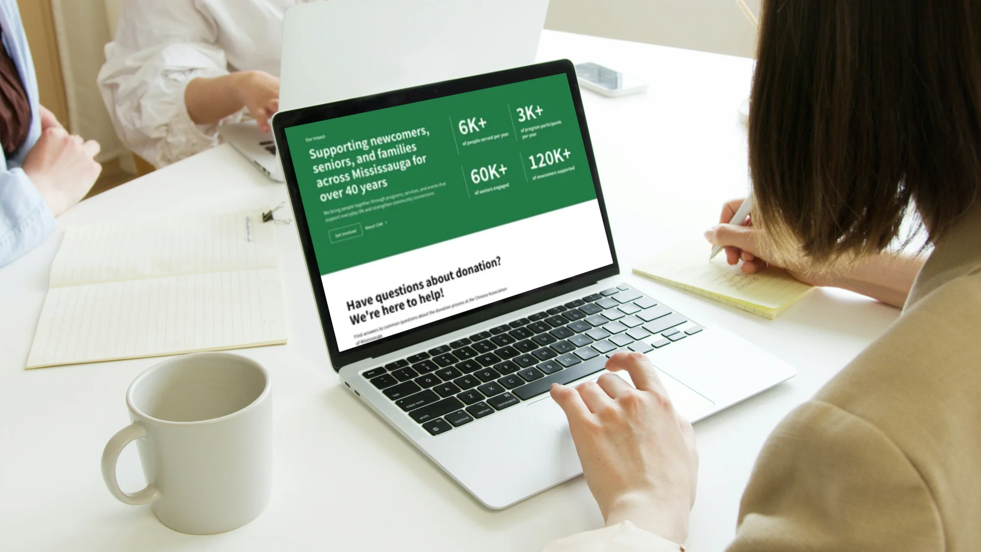 Person using a laptop displaying Chinese Association of Mississauga website about supporting newcomers, seniors, and families across Mississauga with donation information, while another person takes notes beside an empty mug and an open notebook on a white table.
