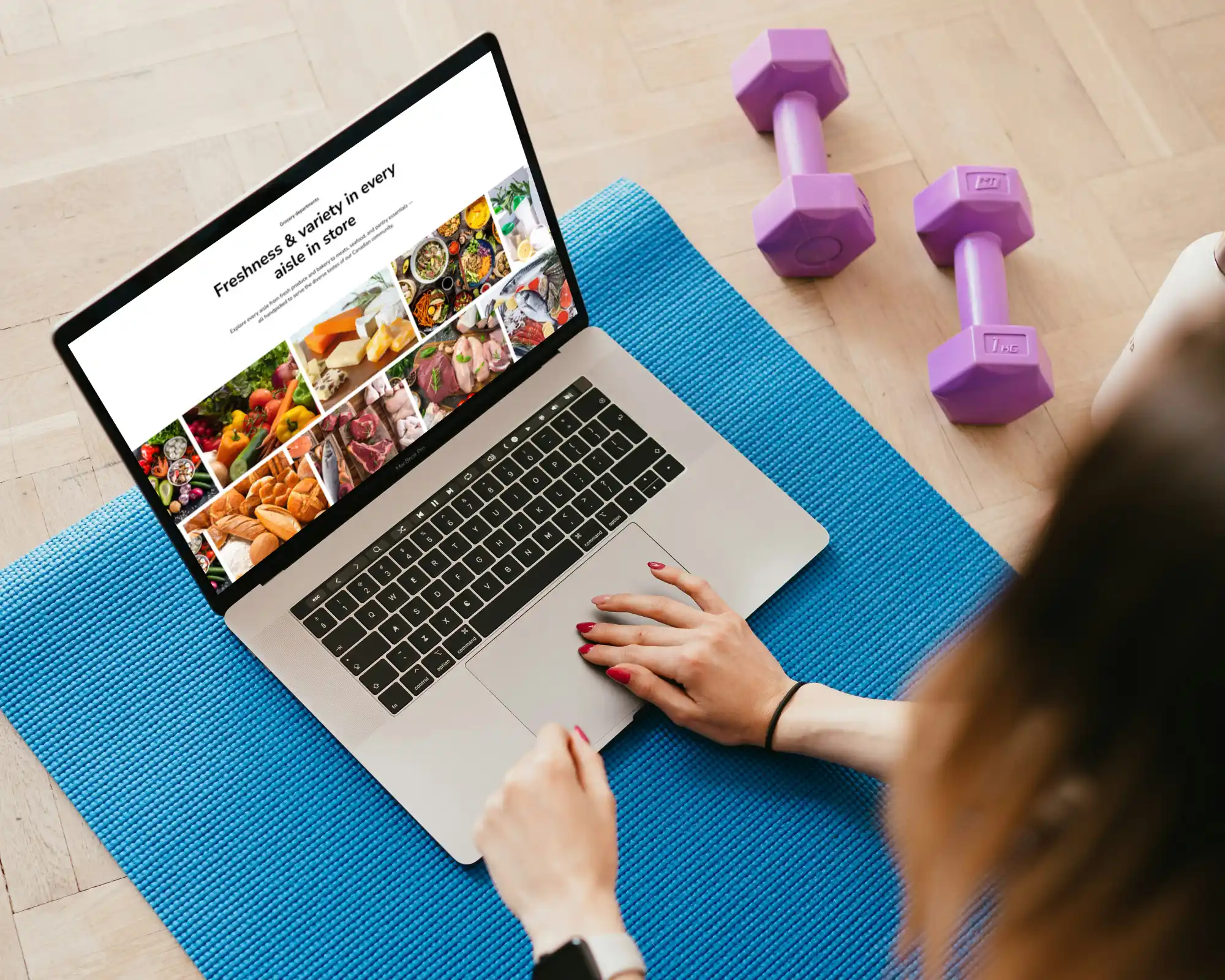 Person viewing JH Supermarket's website on a laptop on a blue exercise mat on a wooden floor, with two purple dumbbells and a water bottle nearby.