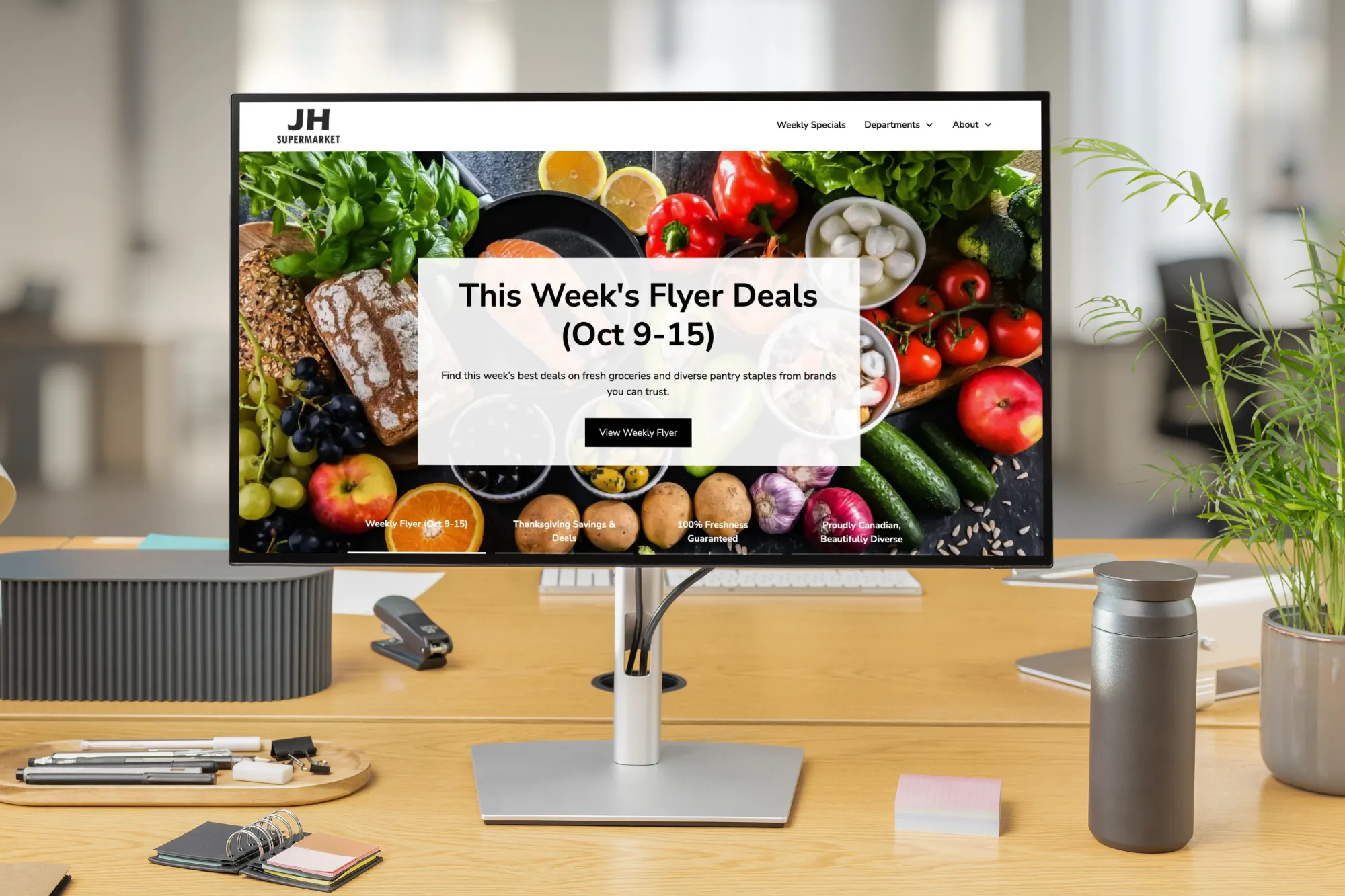 Desktop computer on a wooden desk displaying JH Supermarket website featuring this week's flyer deals (Oct 9-15) with fresh produce background and navigation menu.