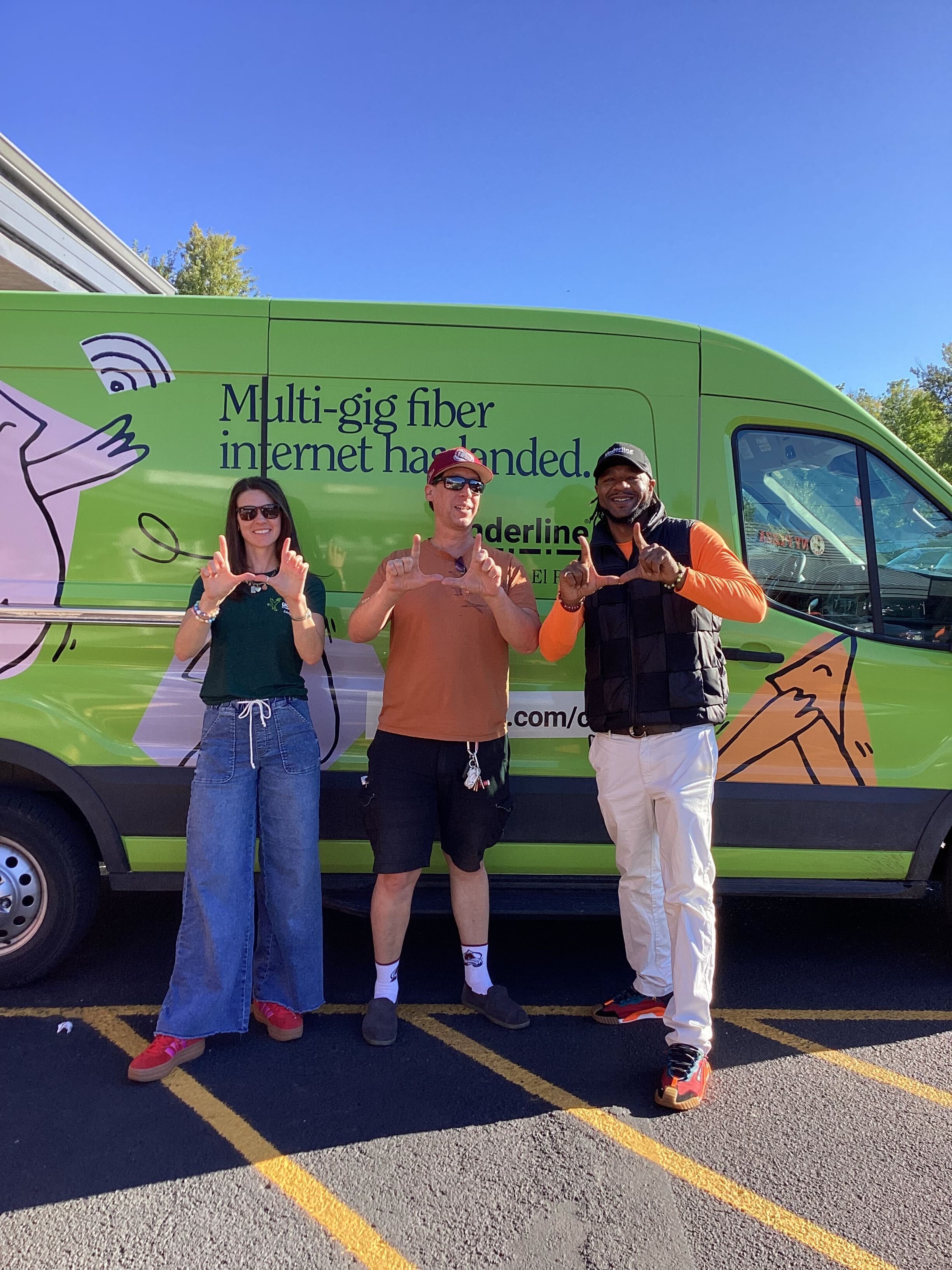 Three people stand in front of a green van with fiber internet ad, making 'L' shapes with their hands.