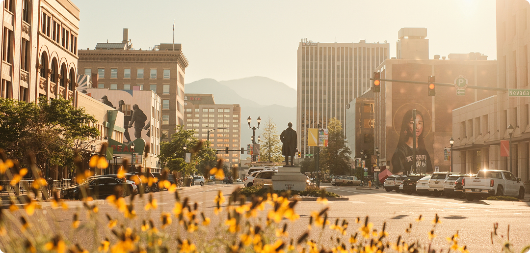 City street at sunset with mountains, yellow flowers, buildings, cars, and a statue in the center.