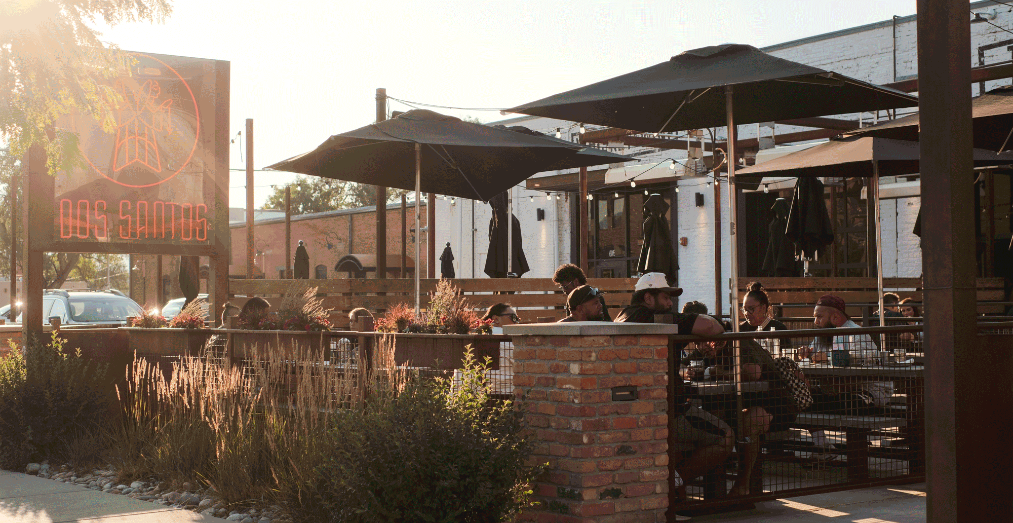 People seated at outdoor tables under black umbrellas at Dos Santos restaurant during sunset.