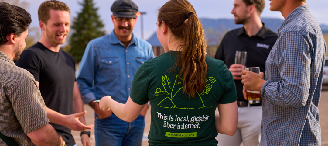 Group of six people talking outdoors; one wears shirt reading 'This is local, gigabit fiber internet.'