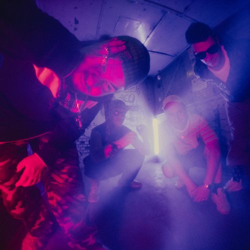 Na zdjęciu widać czterech mężczyzn w starym, opuszczonym budynku. Dwóch nachyla się w kierunku kadru, dwóch kuca. W tle z otwartych drzwi wpada do wnętrza światło, rozświetlając po części ich sylwetki.