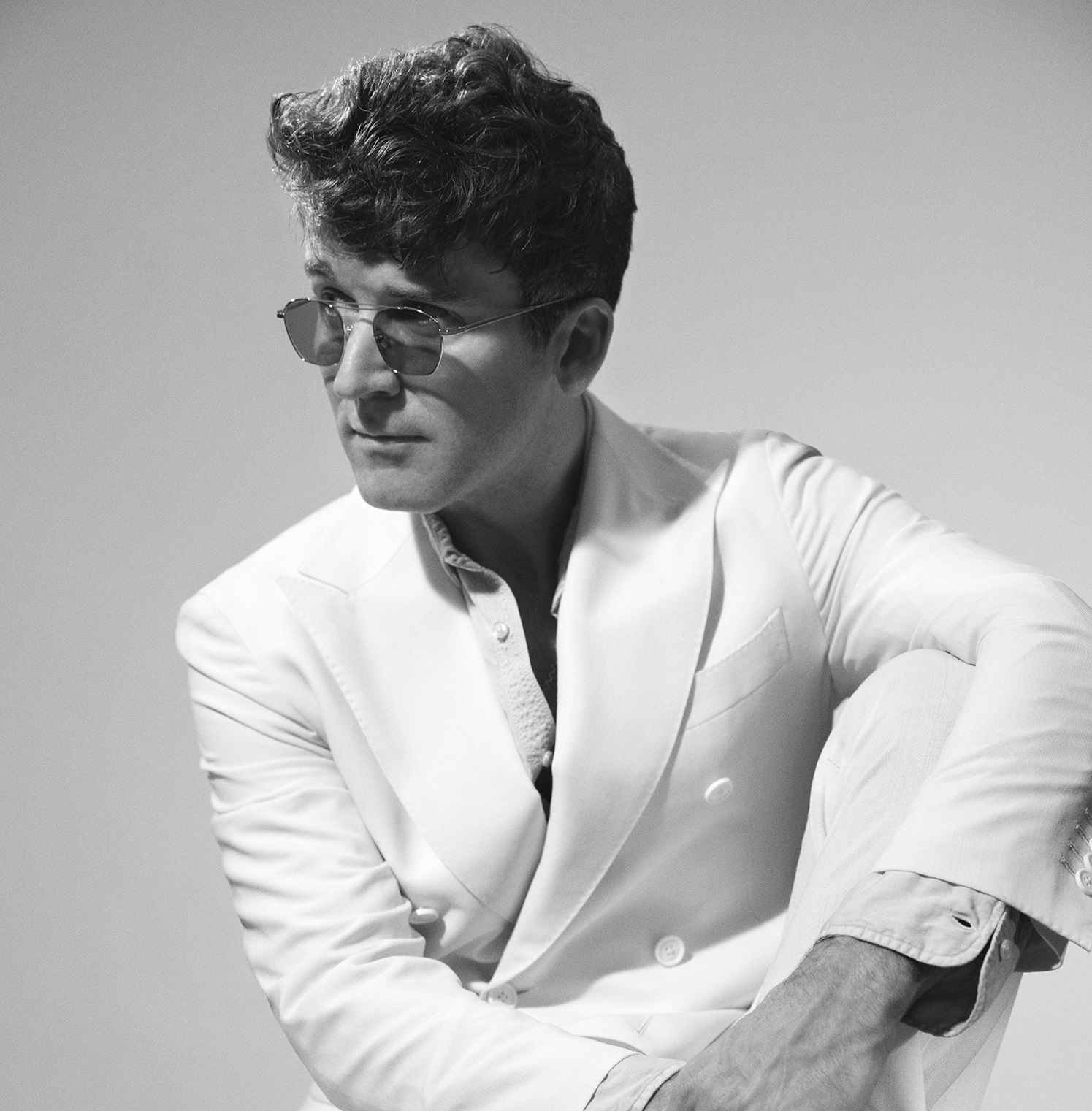 Na zdjęciu widać siedzącego mężczyznę w okularach przeciwsłonecznych w garniturze.